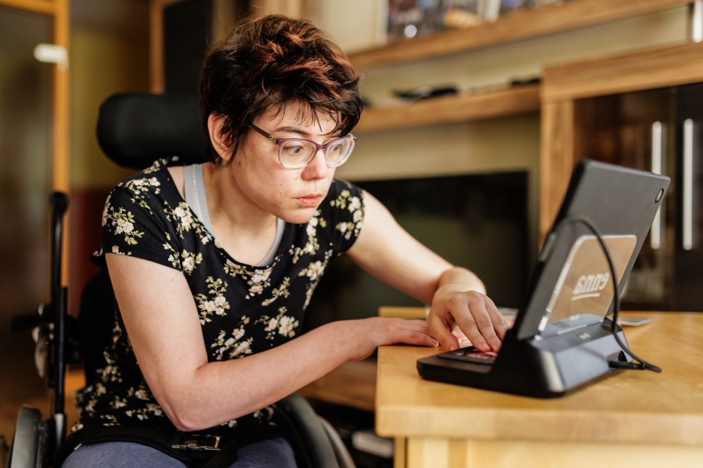 Eine junge Frau sitzt in ihrem Rollstuhl an einem Tisch und blickt auf ein Tablet