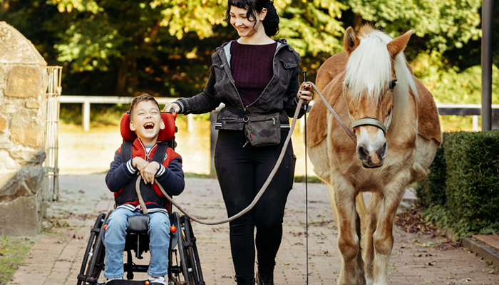 Das Bild zeigt einen Jungen im Rollstuhl mit einer jungen Frau, die ihn beim Führen eines Pferdes begleitet.