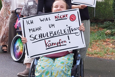 Das Bild zeigt eine junge Frau im Rollstuhl, die ein Schild hochhält mit der Forderung nicht um Schulbegleitung kämpfen zu müssen.